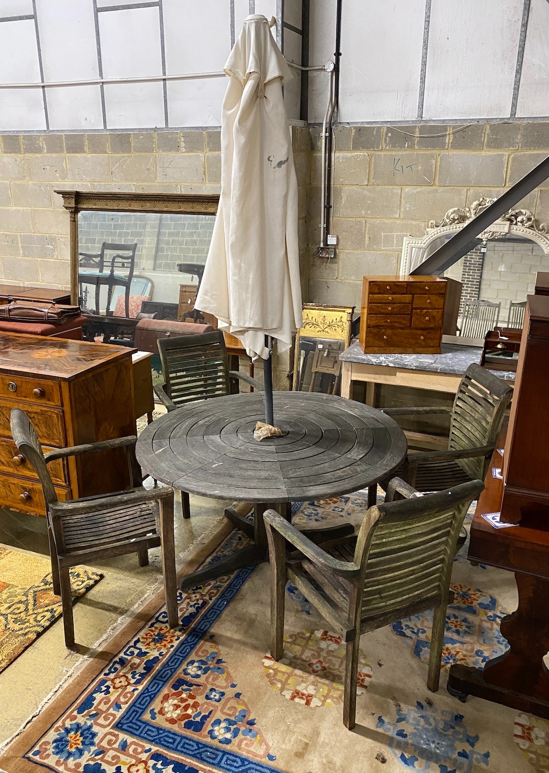 A circular weathered teak garden table, diameter 130cm, height 71cm together with four elbow chairs and a crank parasol